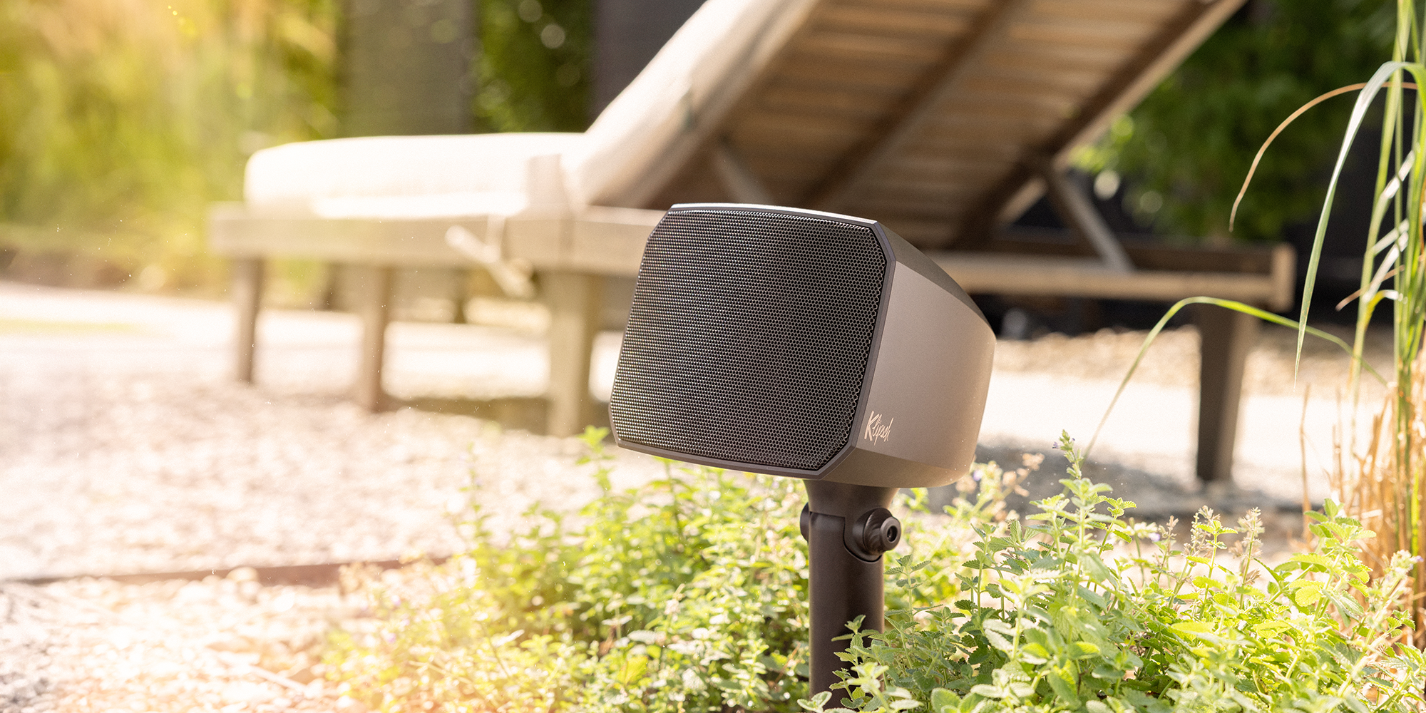 Klipsch Courtyard Satellite Speaker on rock patio up close near lounge chair 2000x750