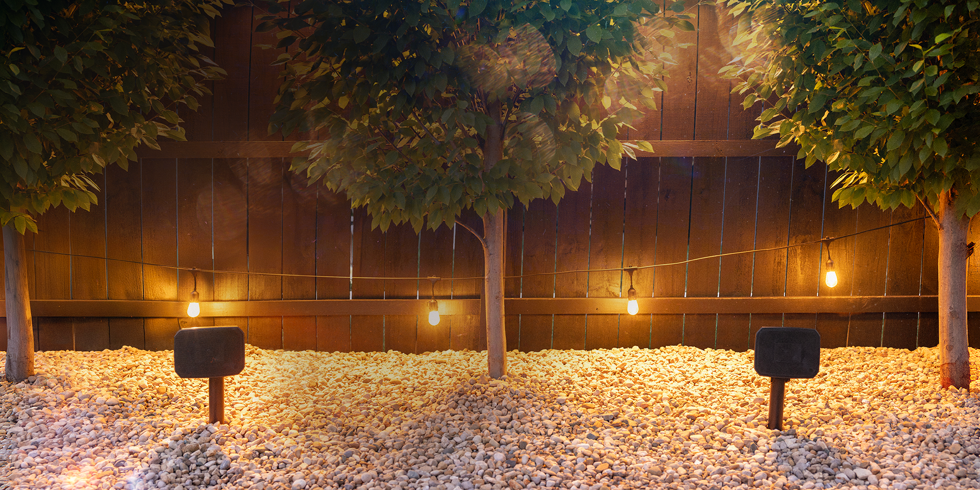 Klipsch Courtyard Speakers outdoors in gravel between symmetrical trees 2 2000x1000