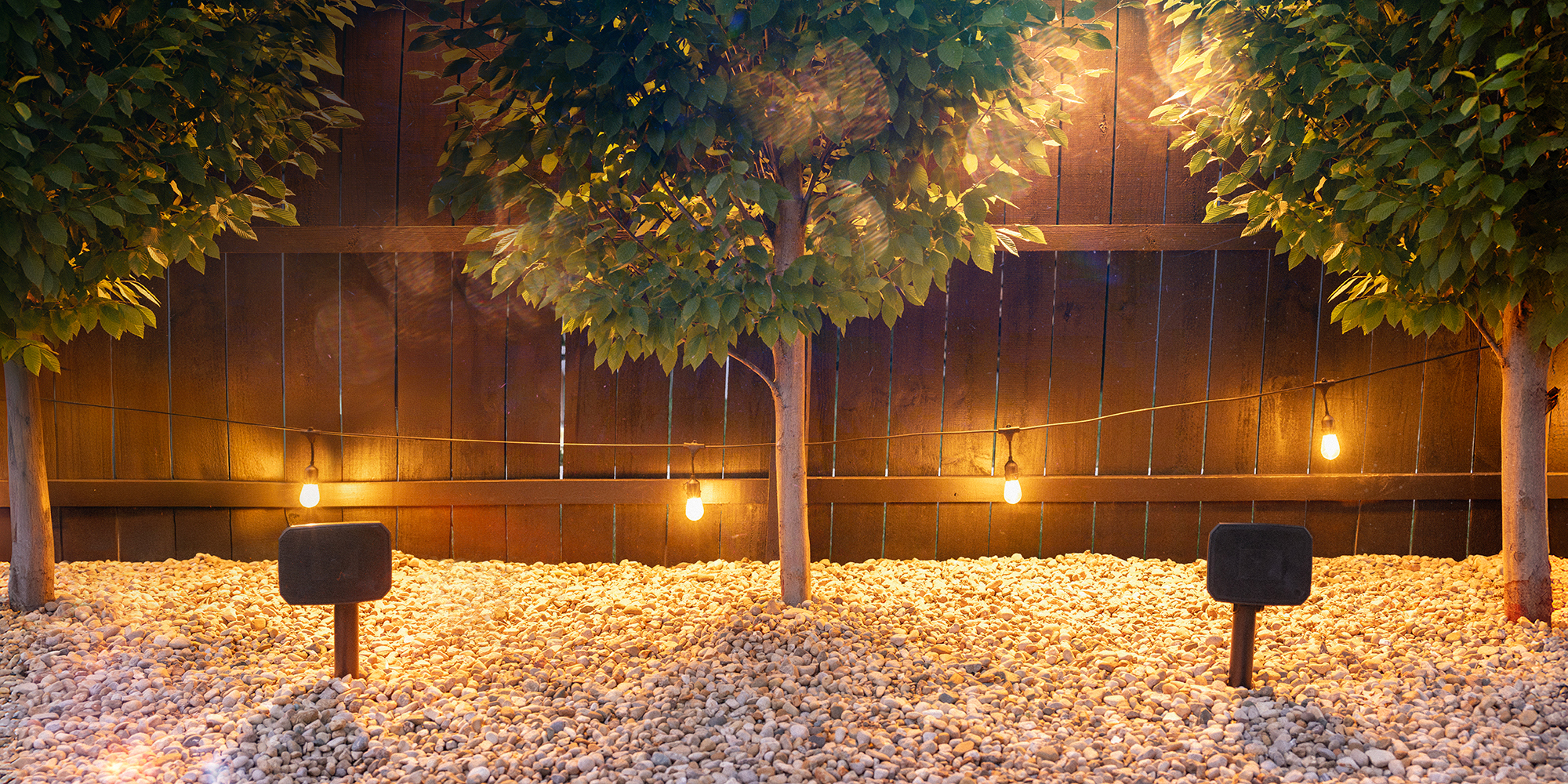 Klipsch Courtyard Speakers outdoors in gravel between symmetrical trees 2000x1000