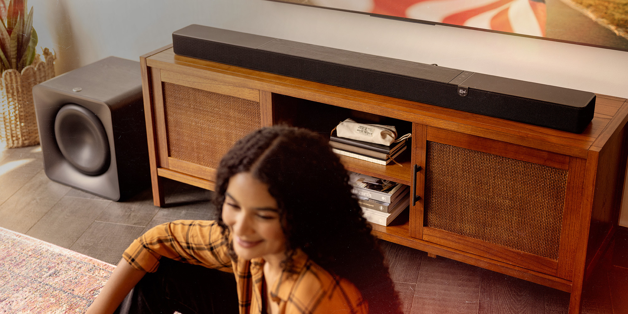 Klipsch Flexus CORE 300 BK and SUB 200 in focus in sunny living room with American flag on tv and woman in blurred foreground overlay 2000x1000