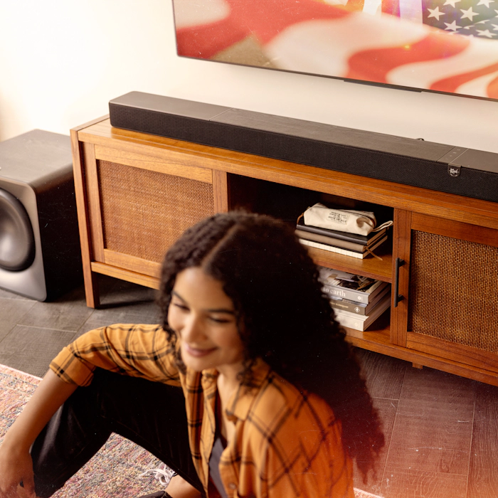 Klipsch Flexus CORE 300 BK and SUB 200 in focus in sunny living room with American flag on tv and woman in blurred foreground 2000x2000
