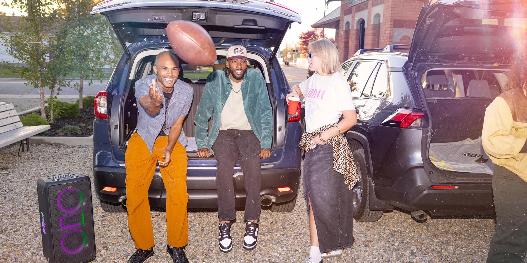 Klipsch Party Speakers Vibe friends at tailgate party playing football and talking behind a car with party speaker nearby in gravel parking lot 2000x1000