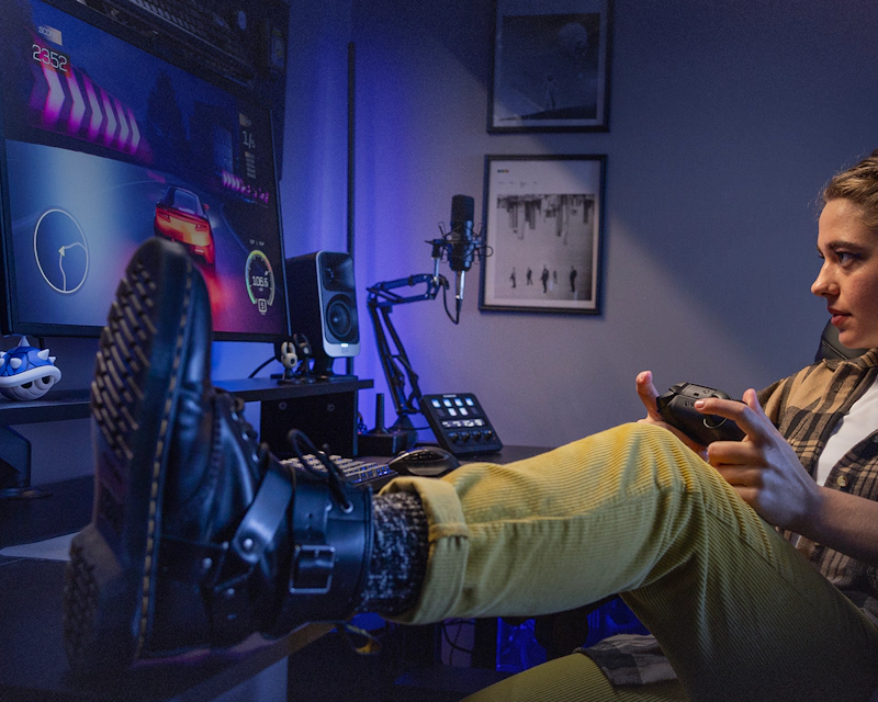 Klipsch Pro Media Lumina Computer Speakers on gamer desk setup with female gamer playing racing game with foot on desk 2000x1000