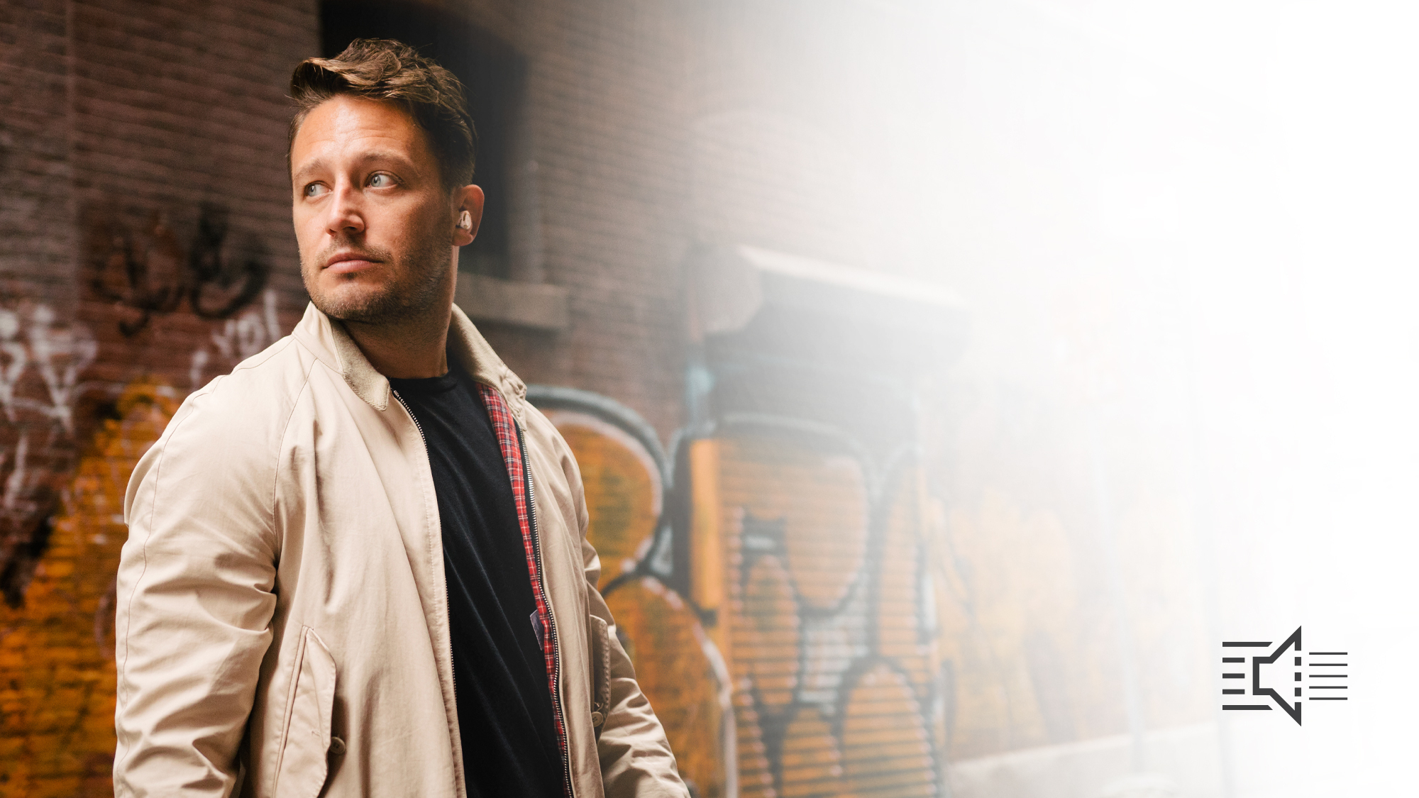 Man standing in front of a graffiti covered brick wall wearing a beige jacket and Klipsch T5 II True Wireless ANC Earphones desktop