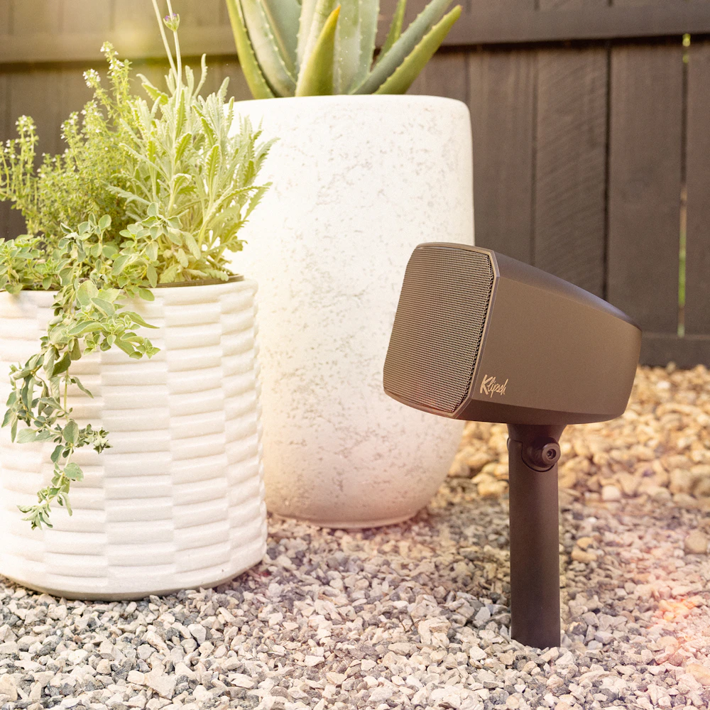 Klipsch Courtyard Speaker outdoors in gravel beside stylish white planters 2000x2000